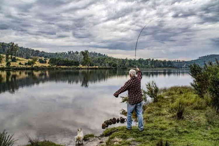 A white-haired man casting a fishing line while standing at the edge of a lake with a small white dog to his side